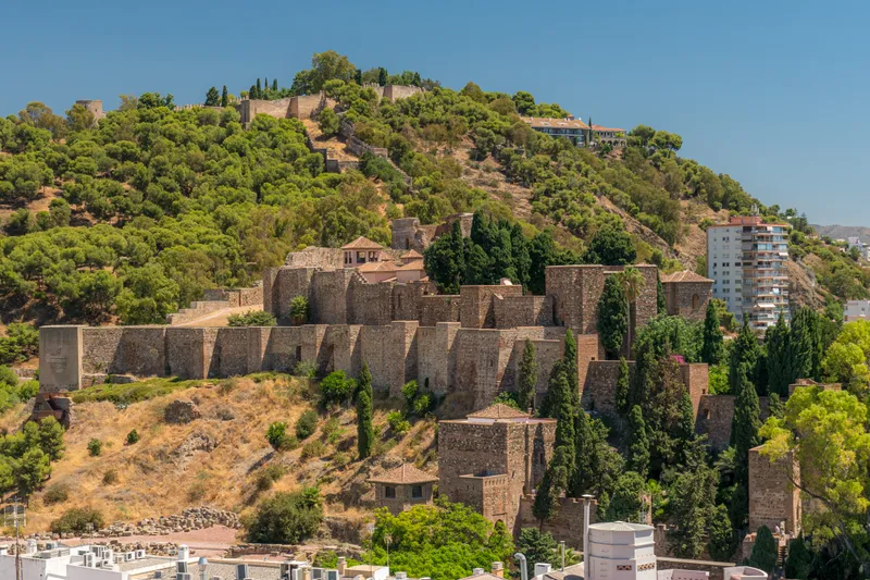 The walls of the Alcazaba climbing the green slope of Gibralfaro hill under a blue sky.